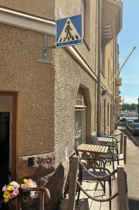 Katunäkymä Encanto Art Cafen ulkopuolelta, ulko-ovi on auki, kuvassa näkyy myös terassia sekä kaukana olevaa merta ja veneitä