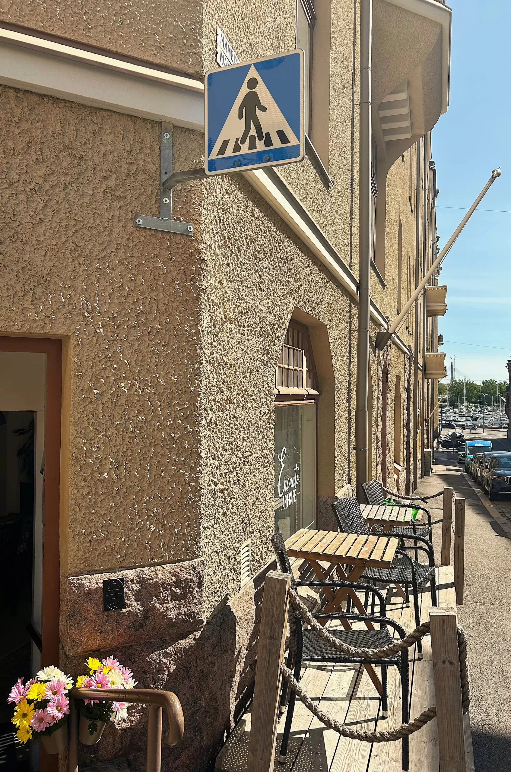 Katunäkymä Encanto Art Cafen ulkopuolelta, ulko-ovi on auki, kuvassa näkyy myös terassia sekä kaukana olevaa merta ja veneitä